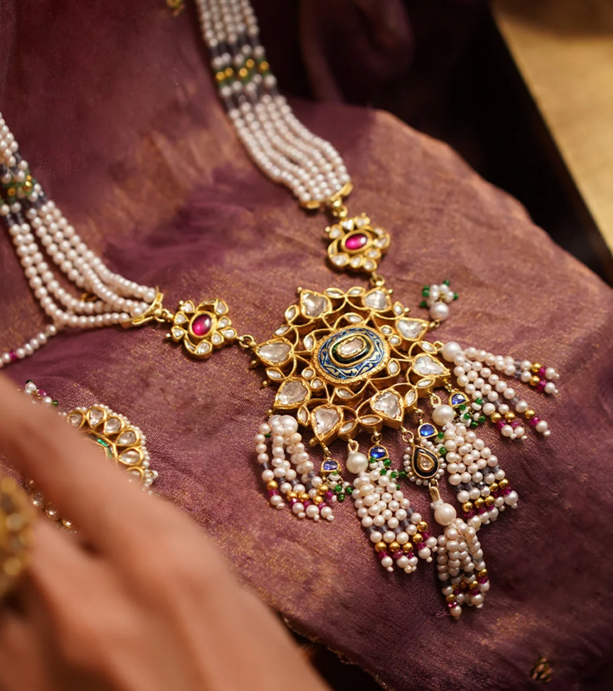 Model wearing handcrafted Yukta gold pendant necklace featuring a six-petalled rosette with kundan-set polki (uncut) diamonds, champlevé meenakari, cultured pearl strands, and pearl tassels accented with iolite, tourmaline beads, and gold balls.
