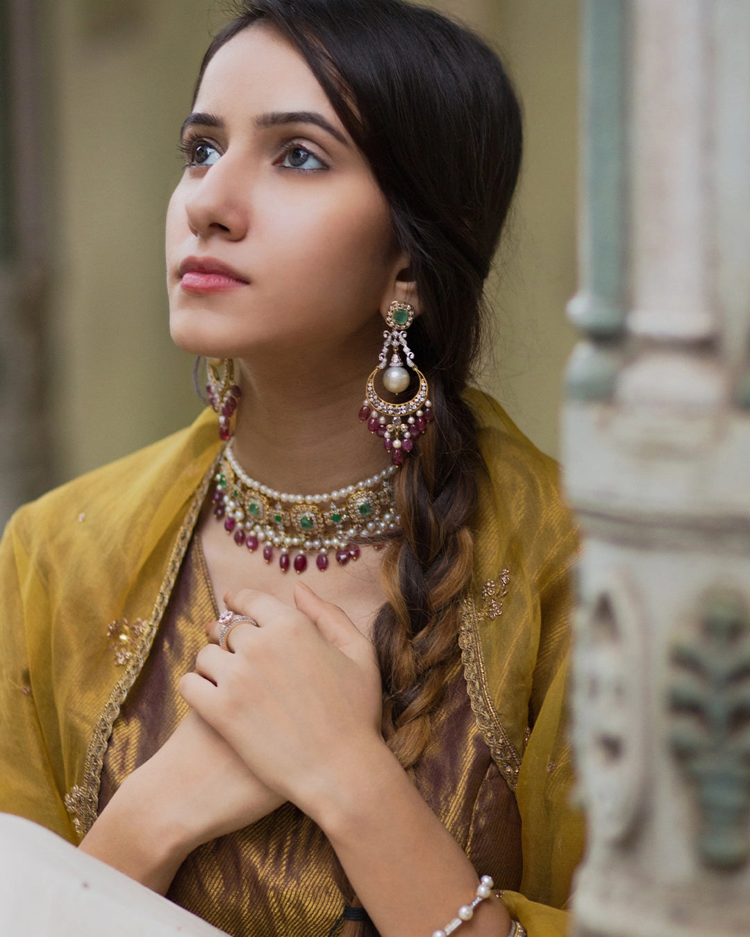 Model wearing the Aurus Jewels Aesha Necklace - a gold necklace with emerald and diamond motifs, pearl strands, and ruby drop fringe, styled as a layered statement neckpiece.